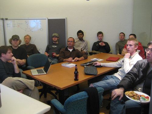A group of eleven men seated around a table, most looking at an unseen screen to the left, one looking at a laptop on the table, another using a wireless keyboard and mouse. Several of them have drinks.