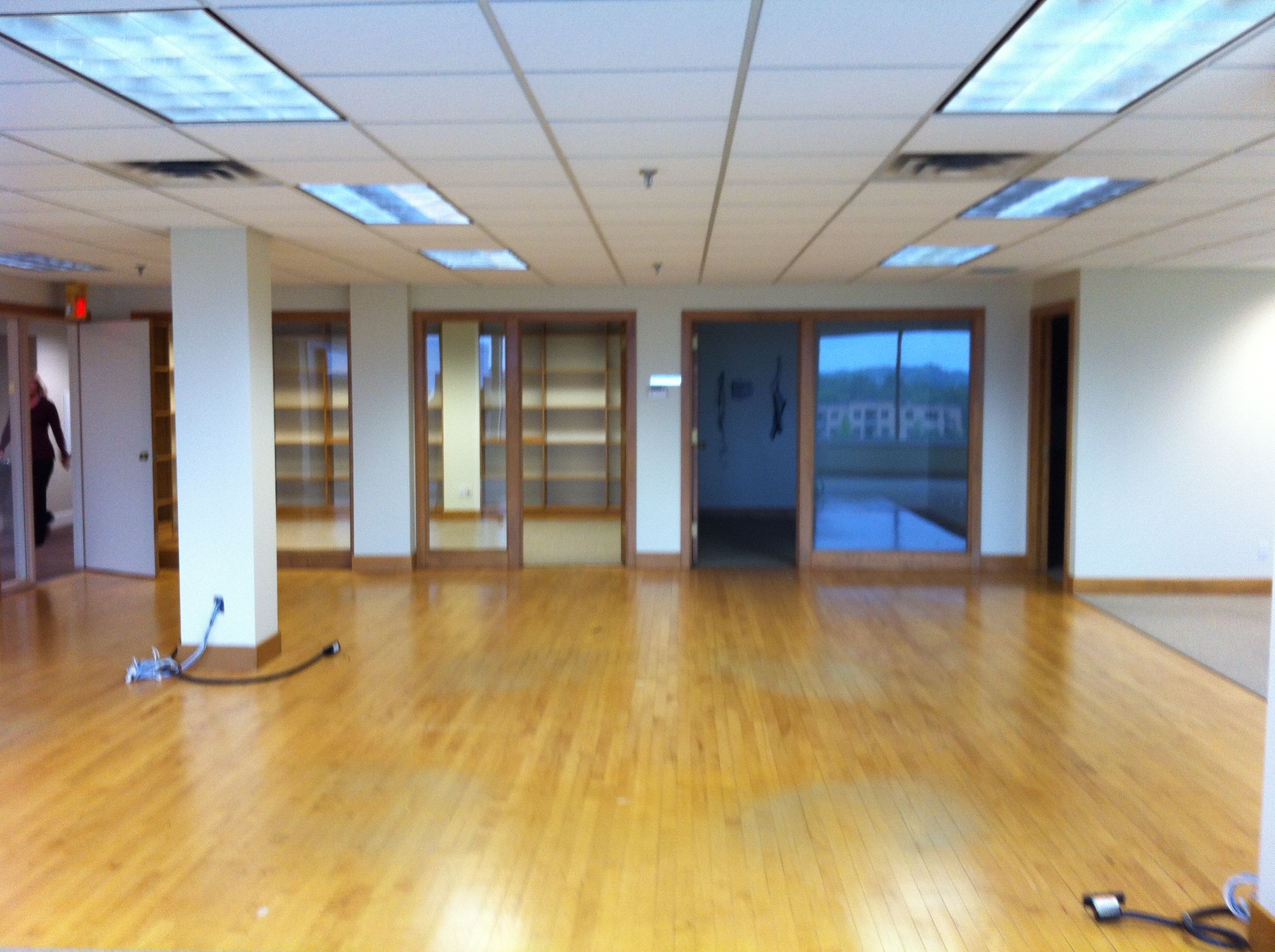 An empty room, most of it has a hardwood floor. A support pillar is visible with some cabling spilling out of an access point. There are smaller rooms at the far end separated room the main one by glass walls. A hallway is visible through a door on the left with a person walking down it.
